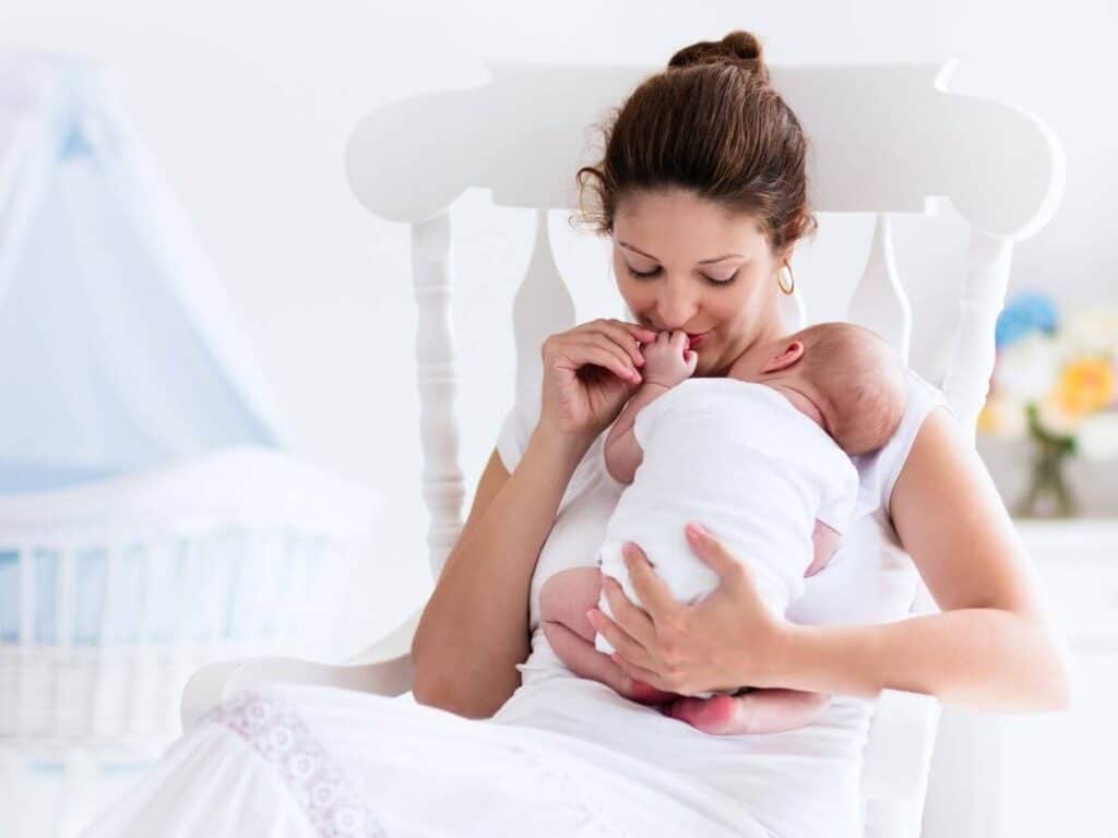 Mom in a rocking chair kissing her newborn baby's hand. How to stop yelling at your kids - remember them as helpless newborns